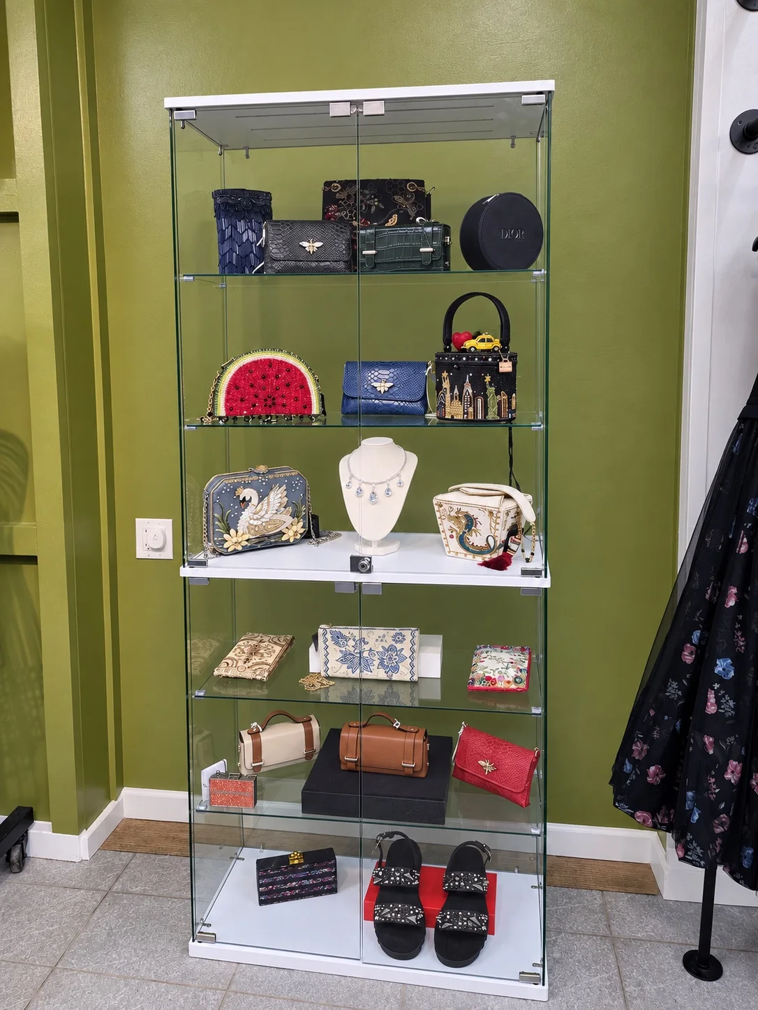 A glass cabinet of handbags, jewelry, and accessories set against an olive wall